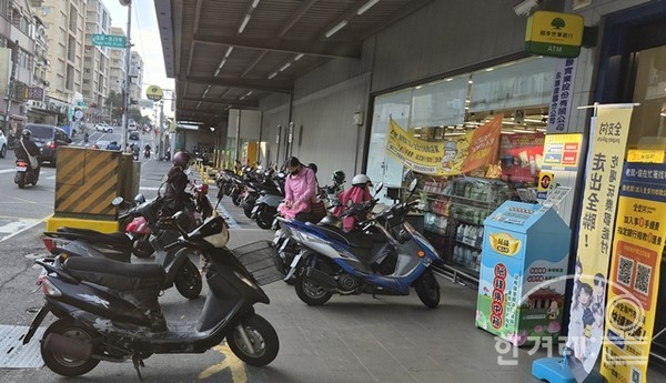편의점에서도 오토바이는 출입구 옆에 좋은 자리. 뒤쪽으로 유료 자동차 주차장. 한 여성이 막 들어오고 있고, 우측 여성은 산 물건을 안장 아래 수납공간에 넣고 있음.